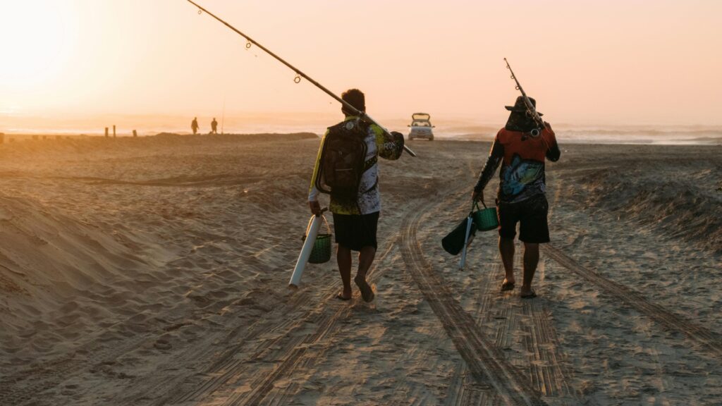 Two men carrying their fishing poles wlaking on the beach at sunset